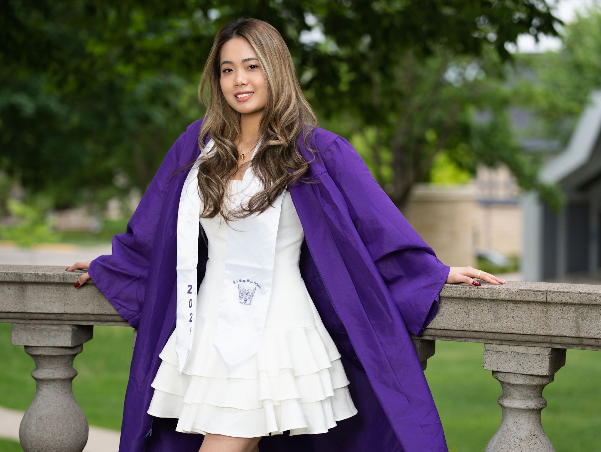 Graduate in a purple gown and white dress leaning on a stone balustrade